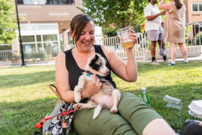 smiling woman holding small puppy and a beer at an outdoor park